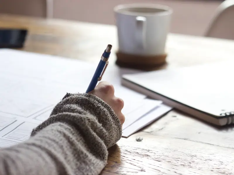 Una mano escribiendo una lista en una libreta, con un café al lado